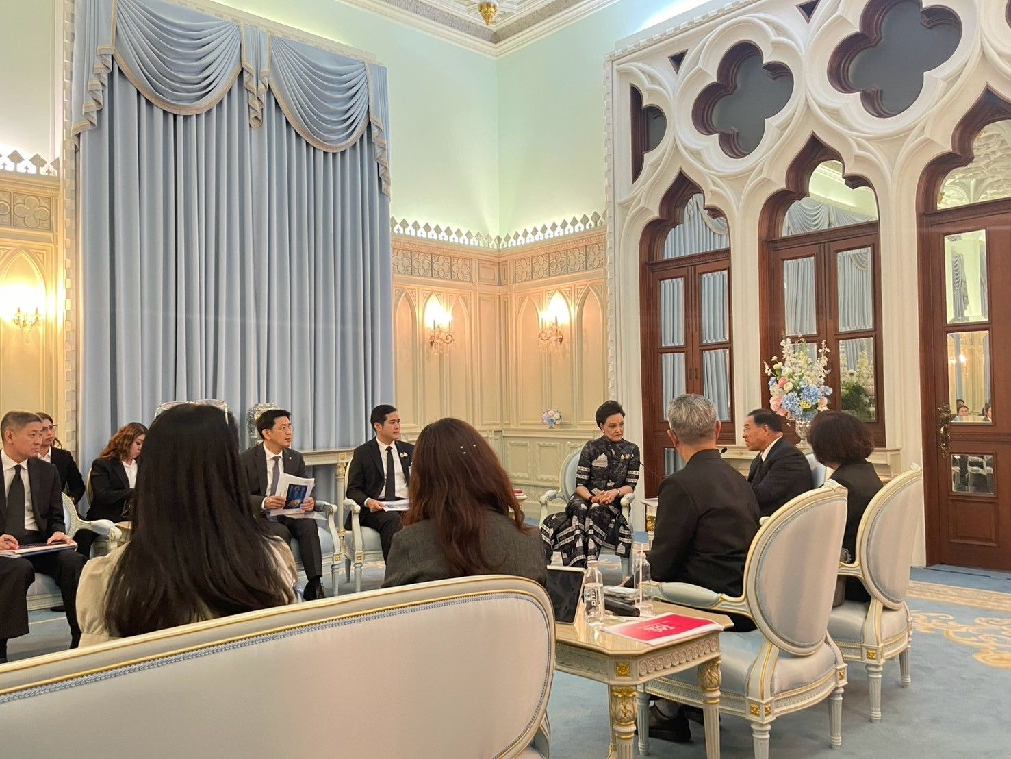 A group of people in business attire are seated in an ornate, formal meeting room.
