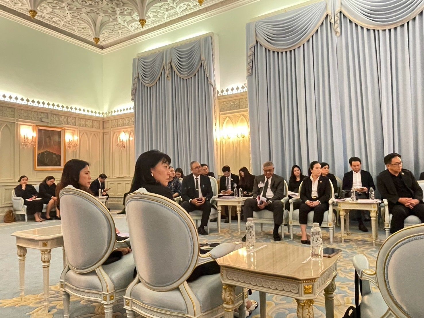 Professionals attend a formal meeting in a lavishly decorated room with high ceilings.