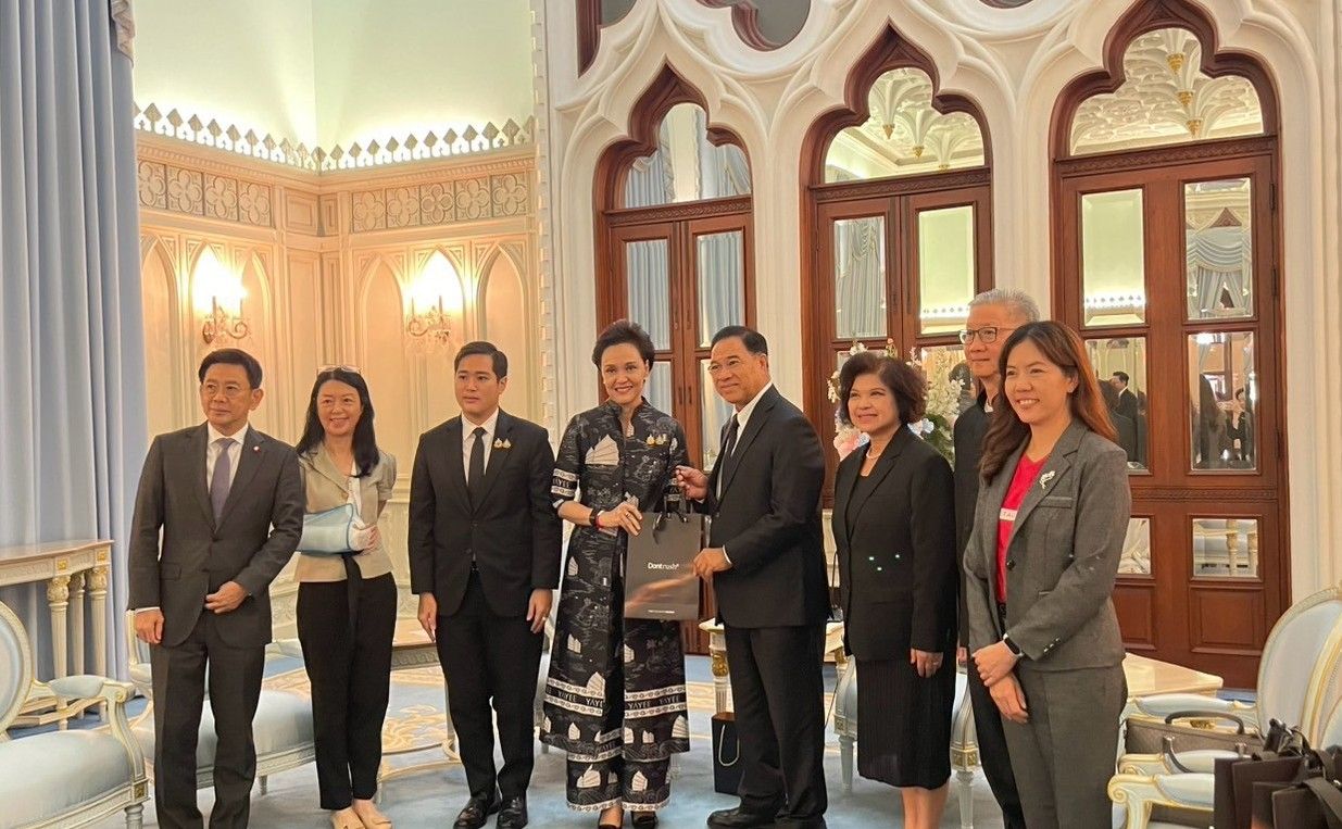 Eight people in formal attire stand in an ornate room, one man gives a gift.