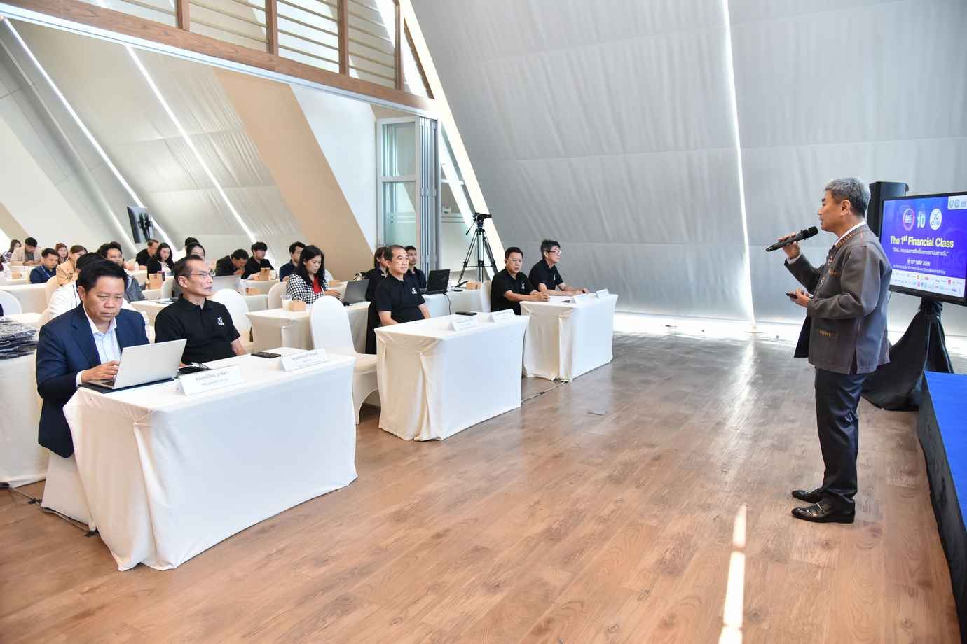 Man presents on stage to an audience seated at tables in a modern conference room.