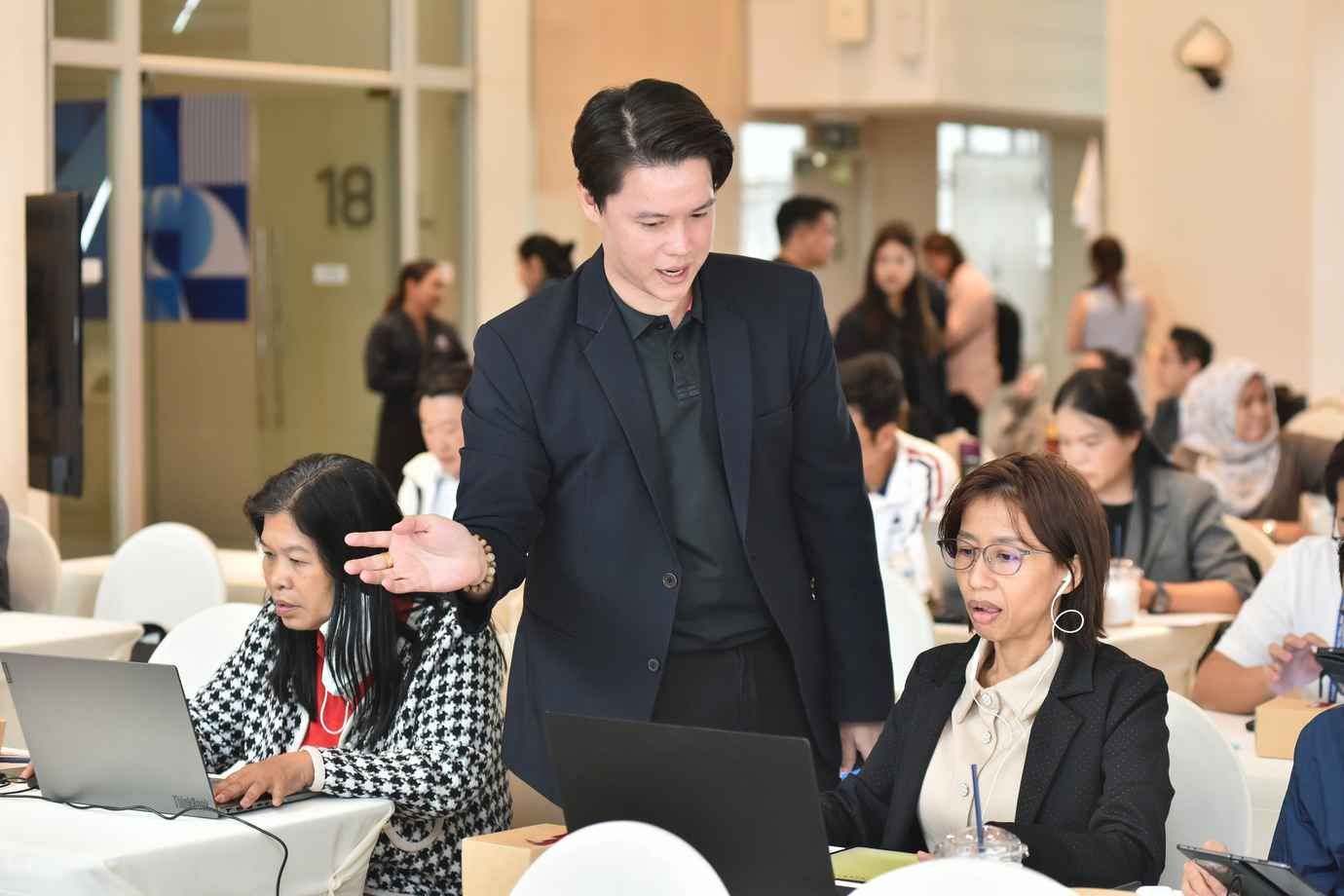 Man in dark suit speaks to women working on laptops at a seminar.
