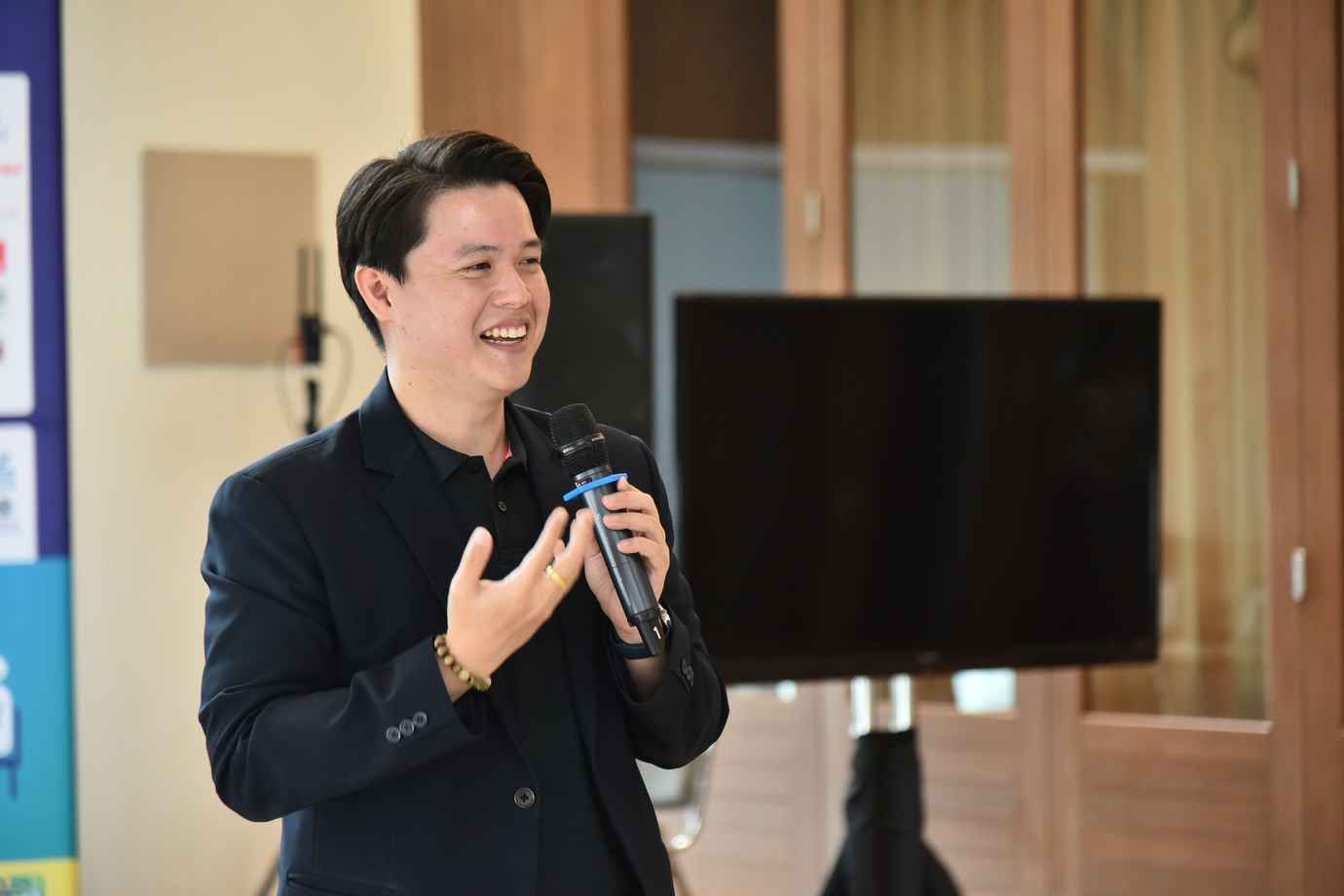 Smiling man in dark suit jacket speaks into microphone, gesturing during a presentation.