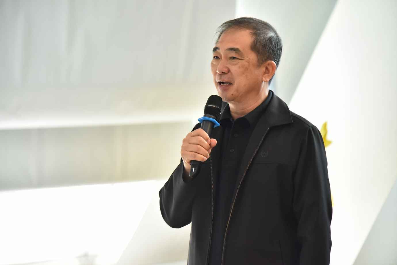 Older Asian man in dark clothes speaks into a microphone.