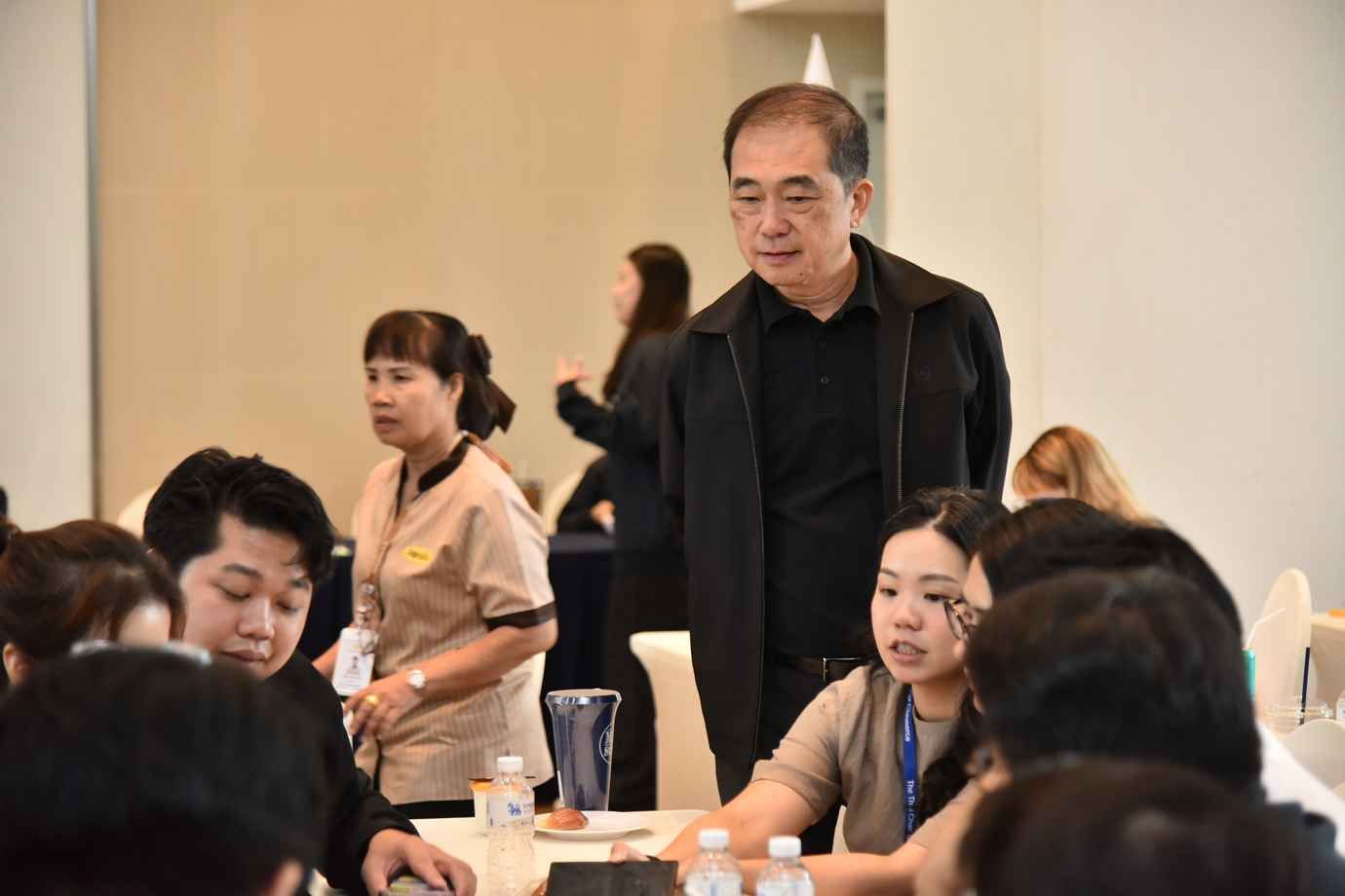 Older Asian man standing, observing seated attendees at an indoor professional event.