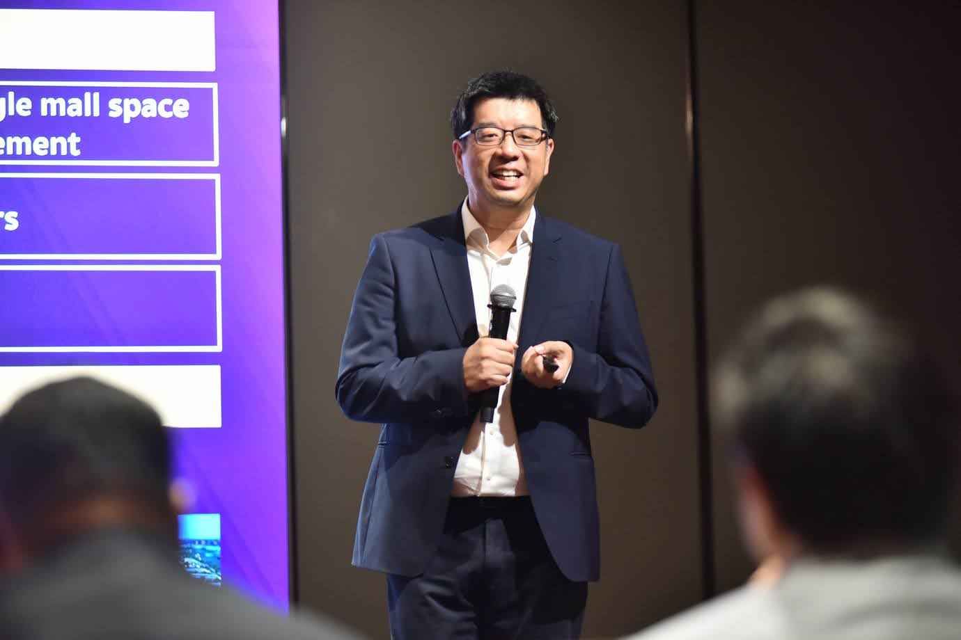 Man in glasses speaks into microphone, presenting in front of a purple screen.