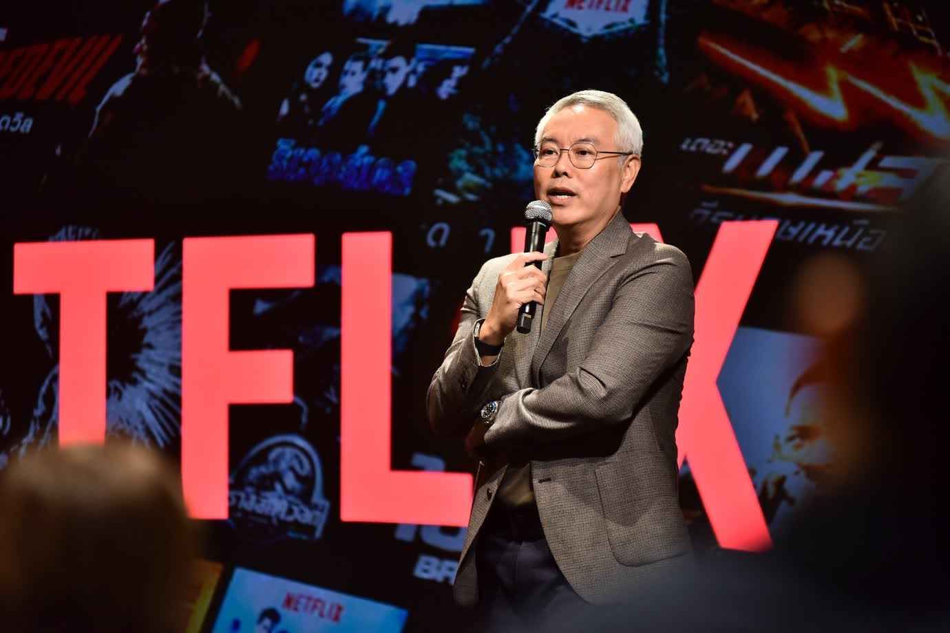 Man in glasses speaking into a microphone in front of a screen displaying 'TFLIX'.