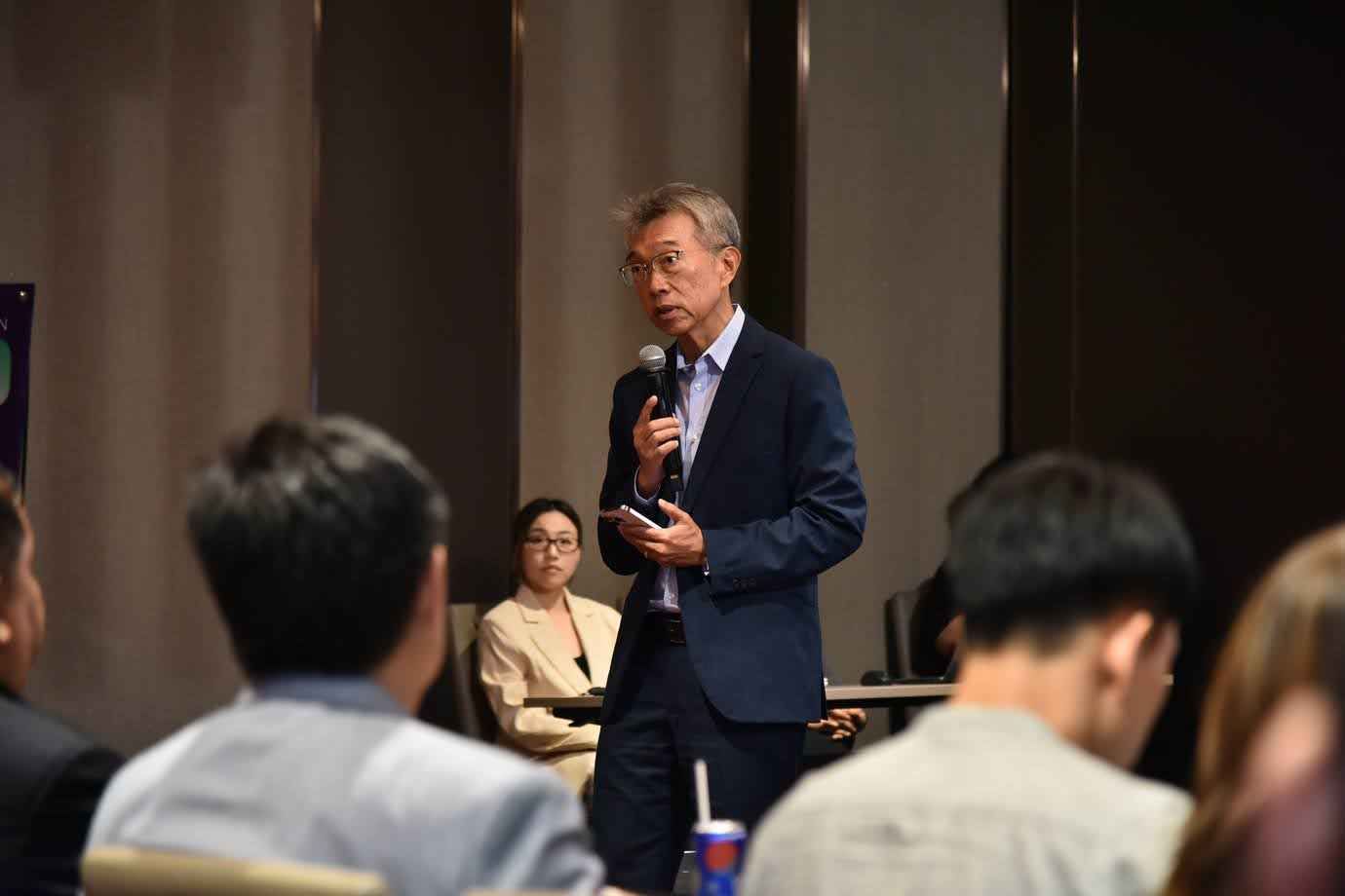 Man in dark suit speaking into a microphone at an event with an audience.
