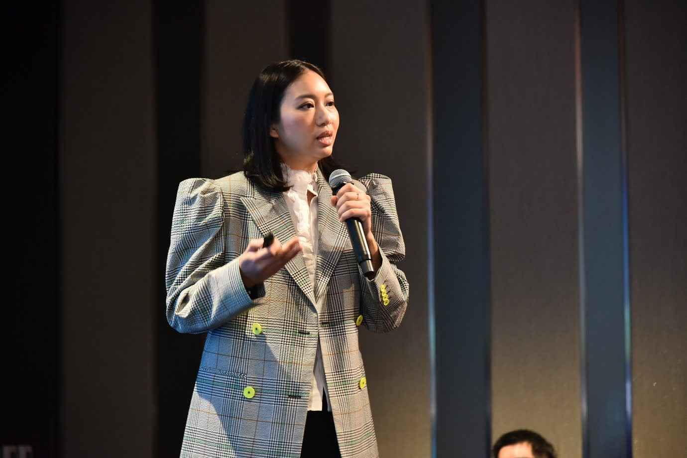 Woman in plaid blazer speaking into microphone, gesturing with hand on stage.