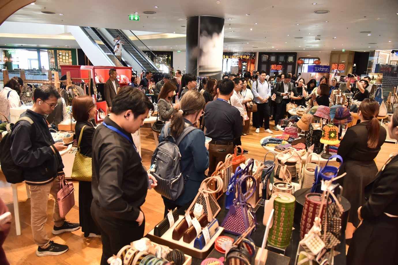 Many shoppers browse a variety of goods at an indoor market with escalators in the background.