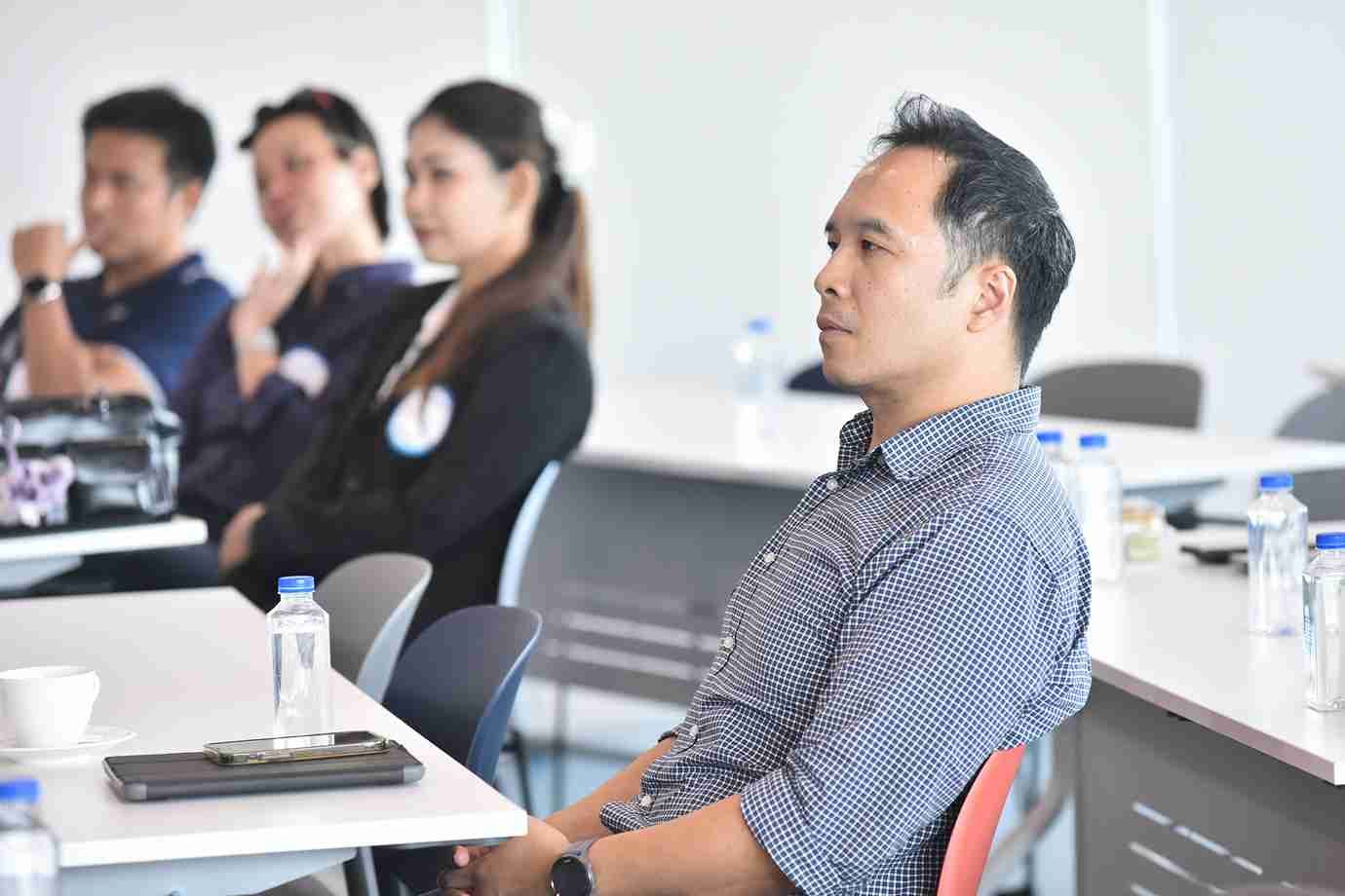 A man in a grey shirt attentively listens during a professional seminar with others in the background.