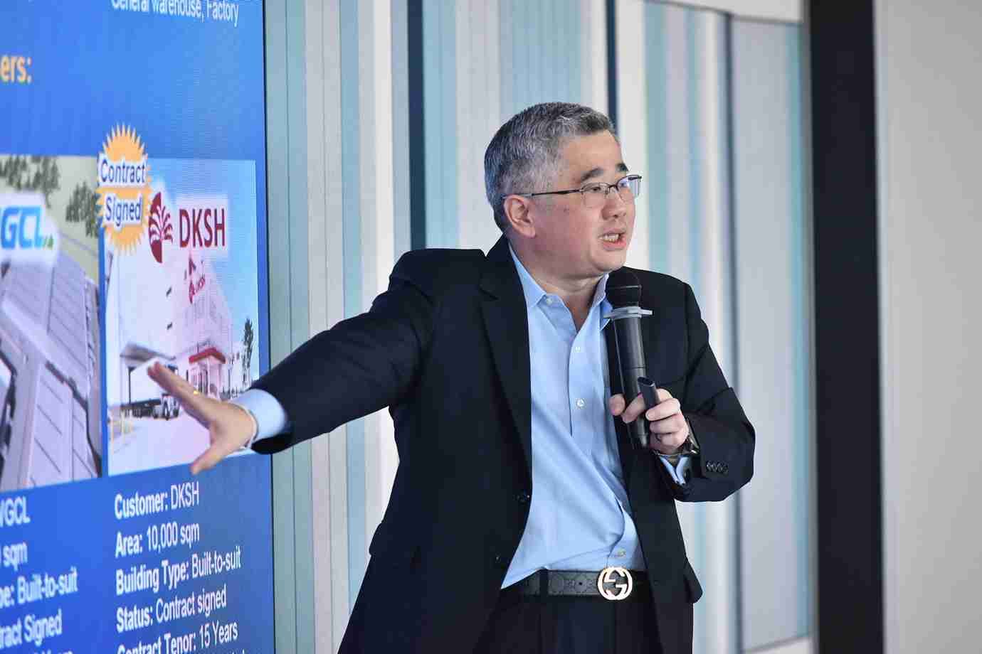 Man in suit speaks into microphone, gesturing towards a presentation screen.