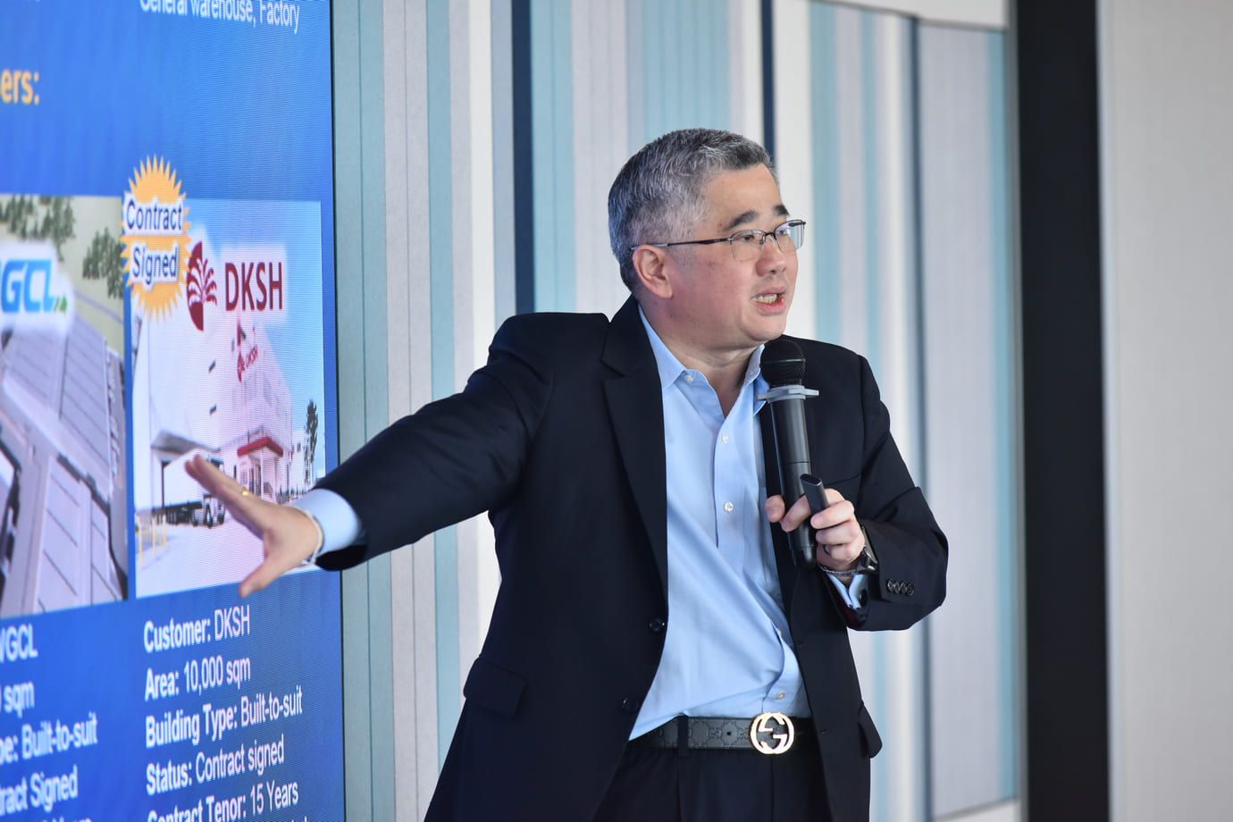A man in a suit speaks into a microphone, gesturing towards a presentation screen.
