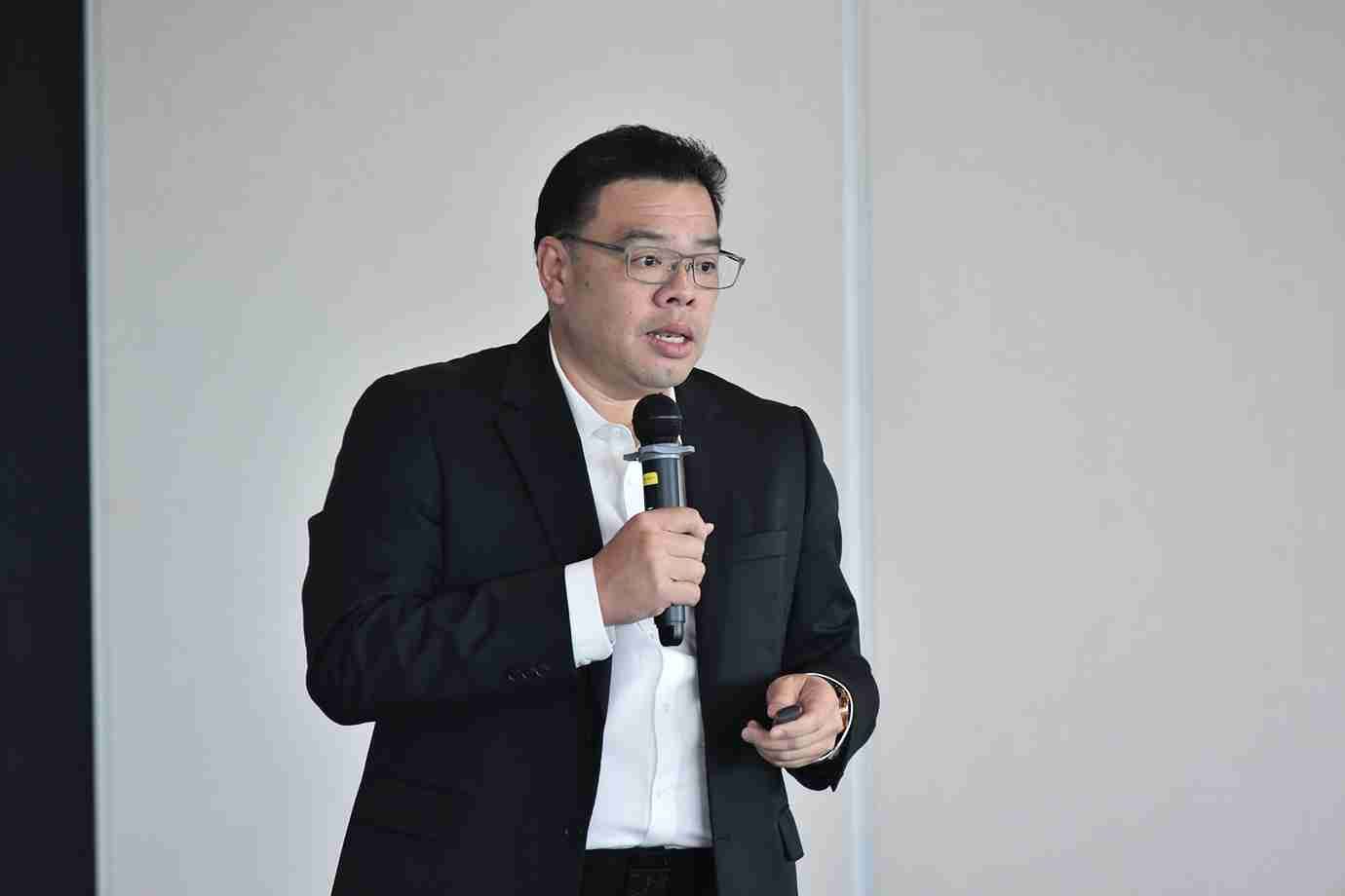 Man in black suit and glasses speaking into a microphone at a presentation.