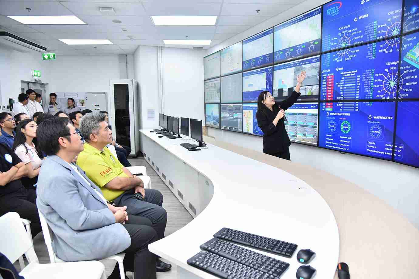 A woman points to complex data on large blue screens as a group of people watch intently.