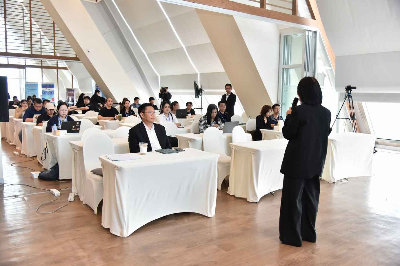 A woman in black suit speaks to a seated audience at tables in a bright conference room.