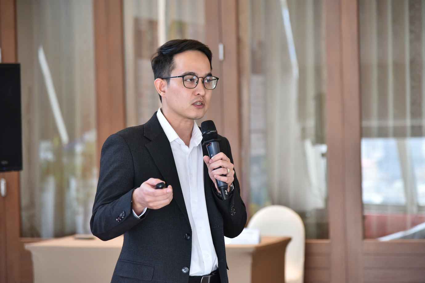 A man in a suit and glasses speaks into a microphone while holding a presentation clicker.