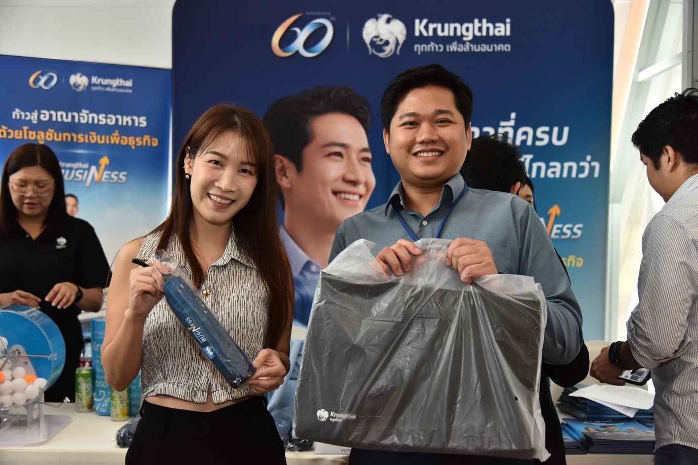 A smiling woman and man hold promotional items in front of a Krungthai Bank backdrop.