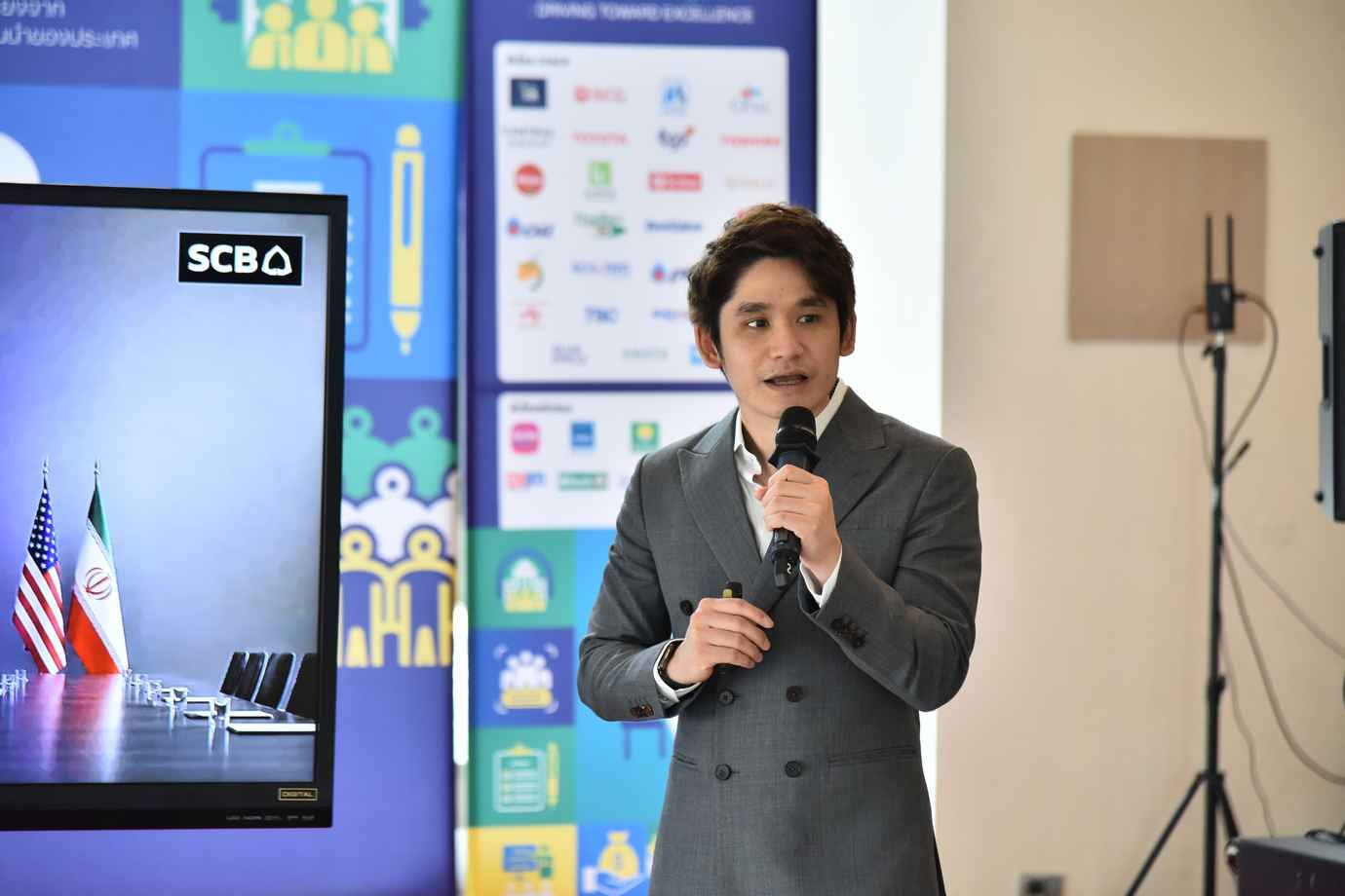 Young man in a gray suit speaks into a microphone during a presentation with a screen.