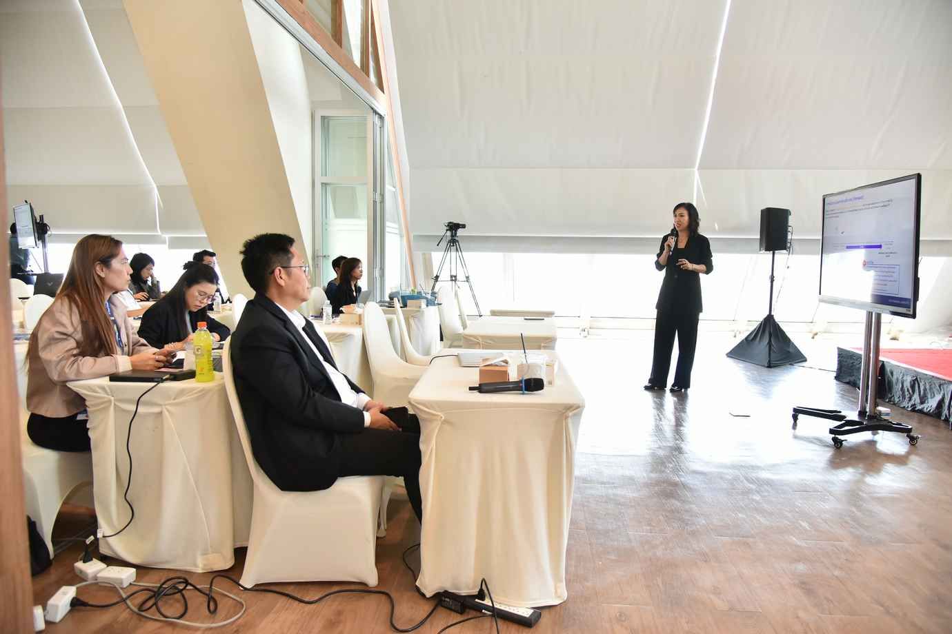 A woman presents to an audience with a large screen in a bright conference room.