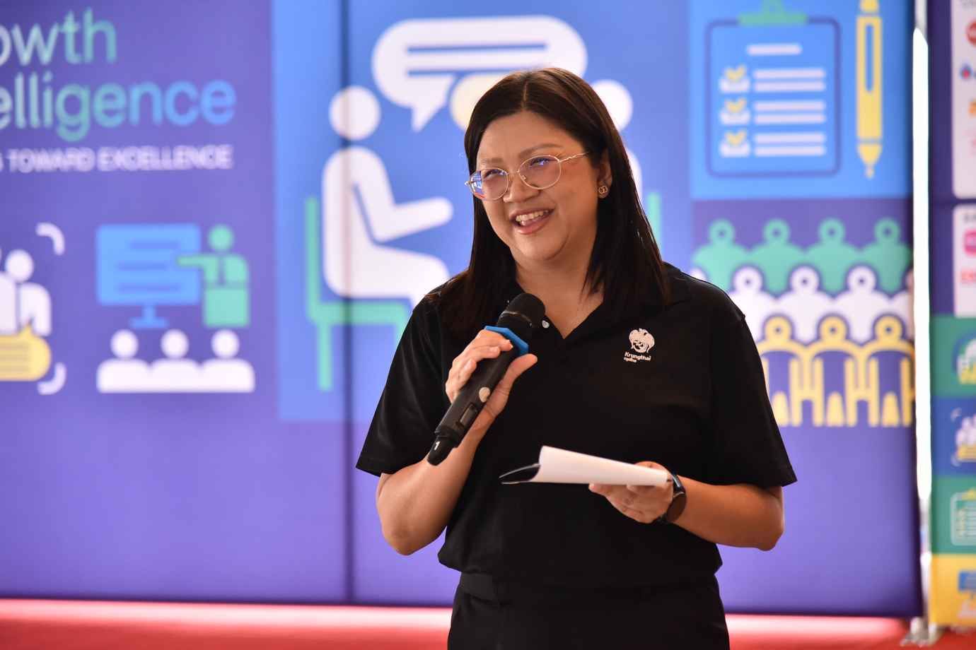 Smiling woman in glasses and black polo speaks into a microphone at a presentation.