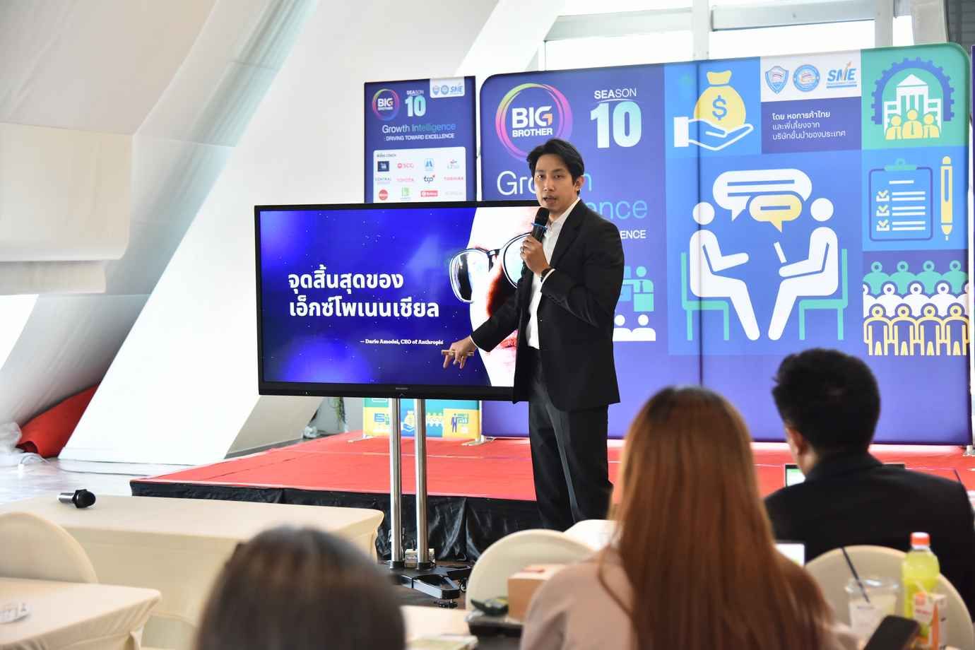 A presenter points at a screen displaying Thai text and graphics to an audience.