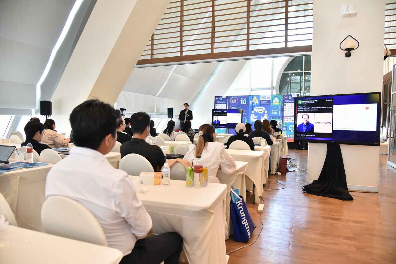 Attendees watch a speaker present at a modern conference, with a large screen displaying content.