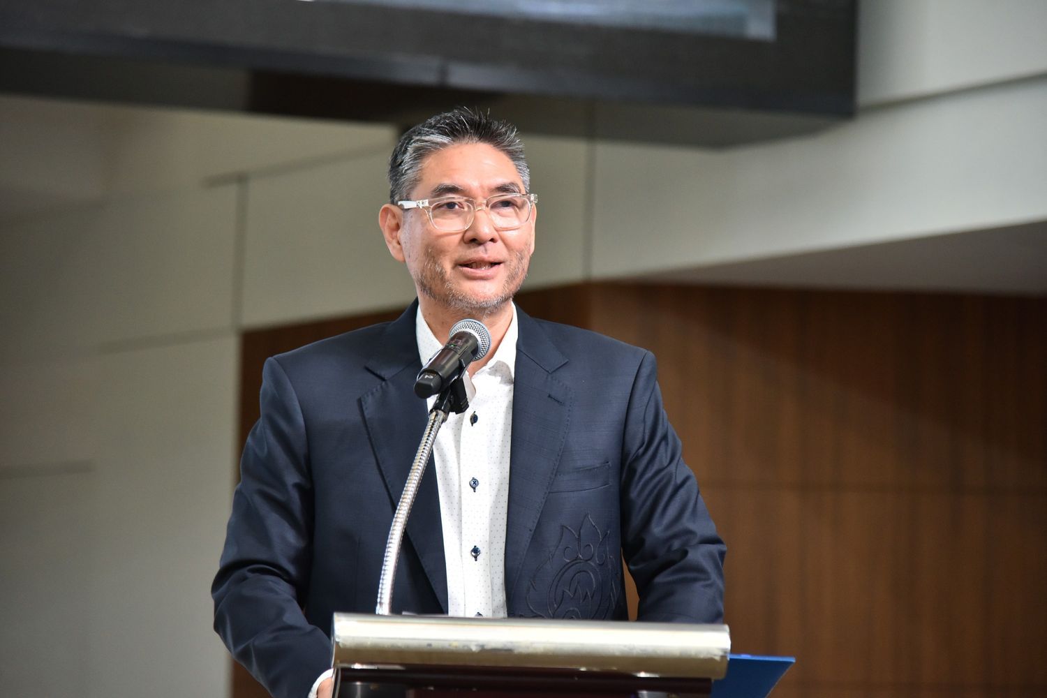 A distinguished man in a dark suit smiles while speaking at a podium with a microphone.