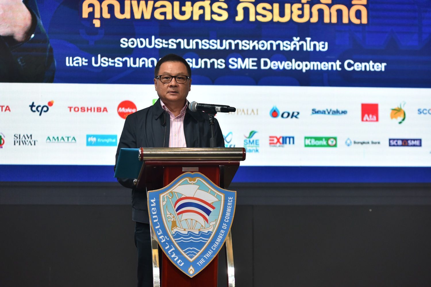 A man speaks at a podium in front of a screen displaying event sponsors' logos and text.