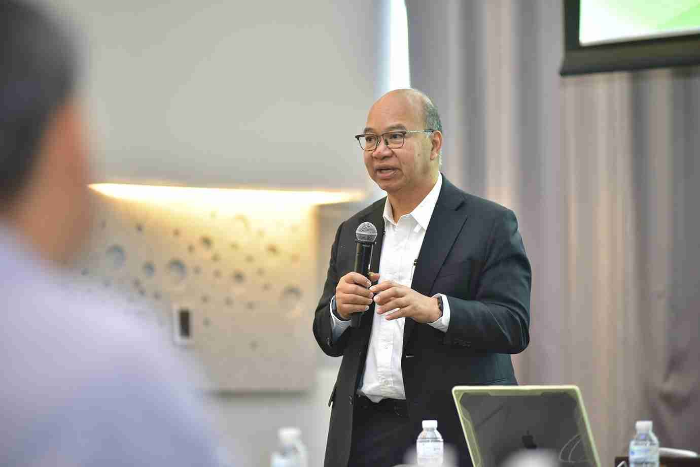 A man in a suit and glasses speaks into a microphone during a presentation.