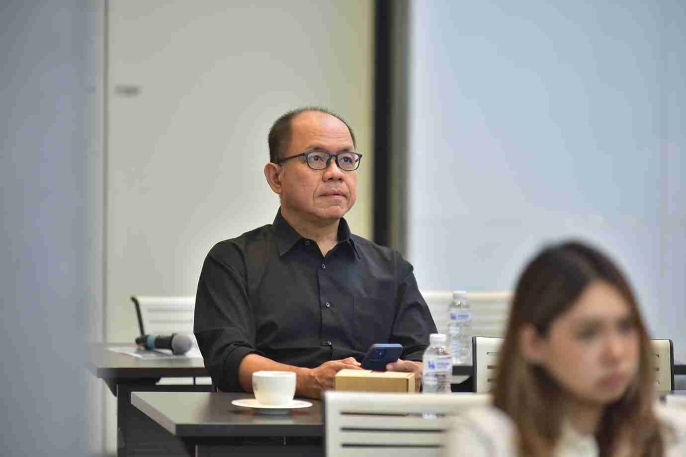 A man in glasses and a black shirt intently observes a presentation in a meeting room.