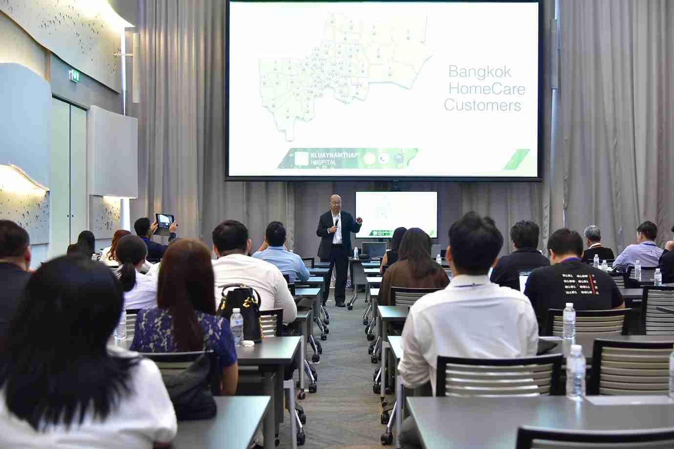 Man presenting to an audience in a conference room with "Bangkok HomeCare Customers" on screen.