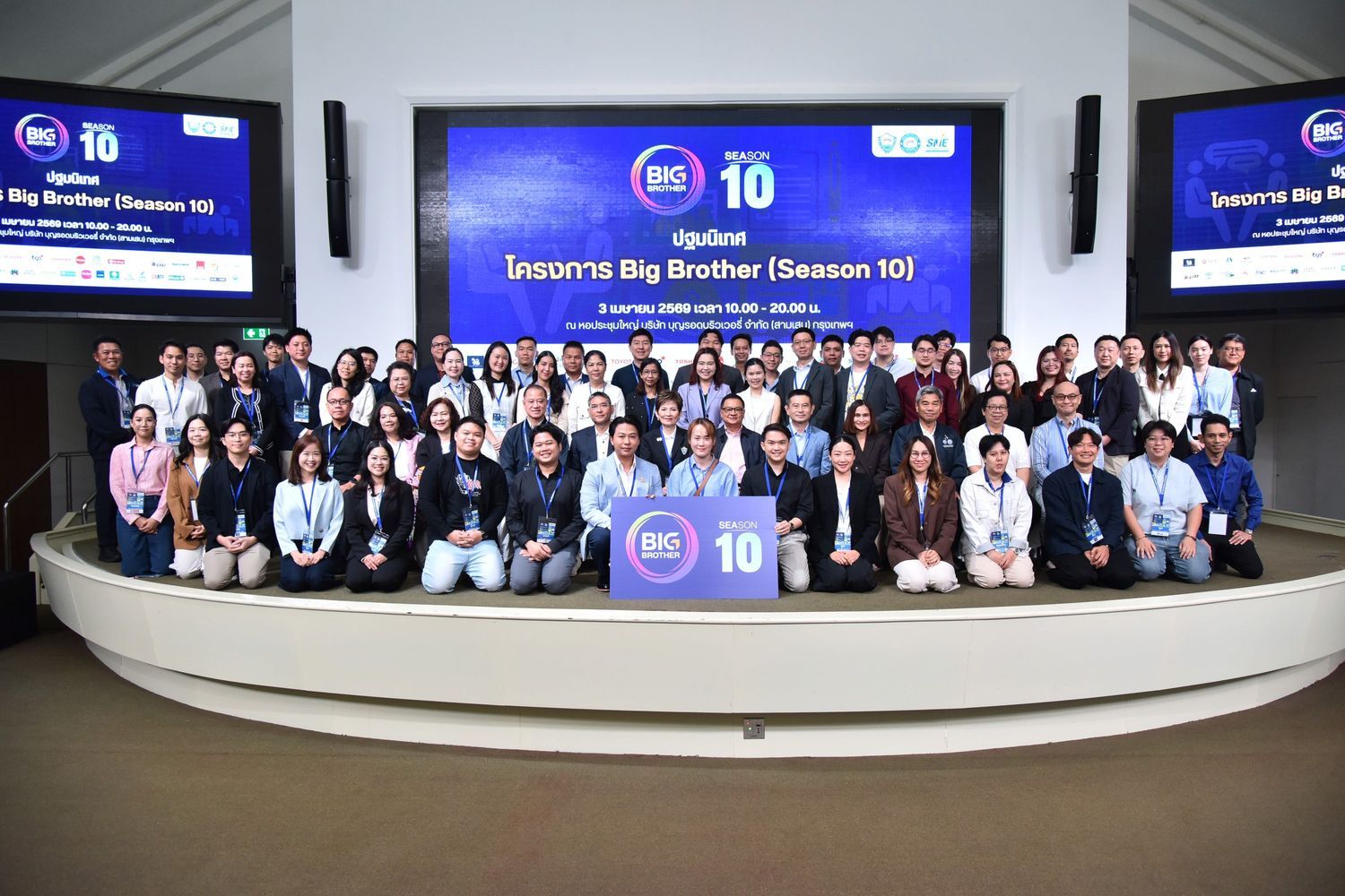 A large group of people pose for a photo in front of two big screens with event branding.