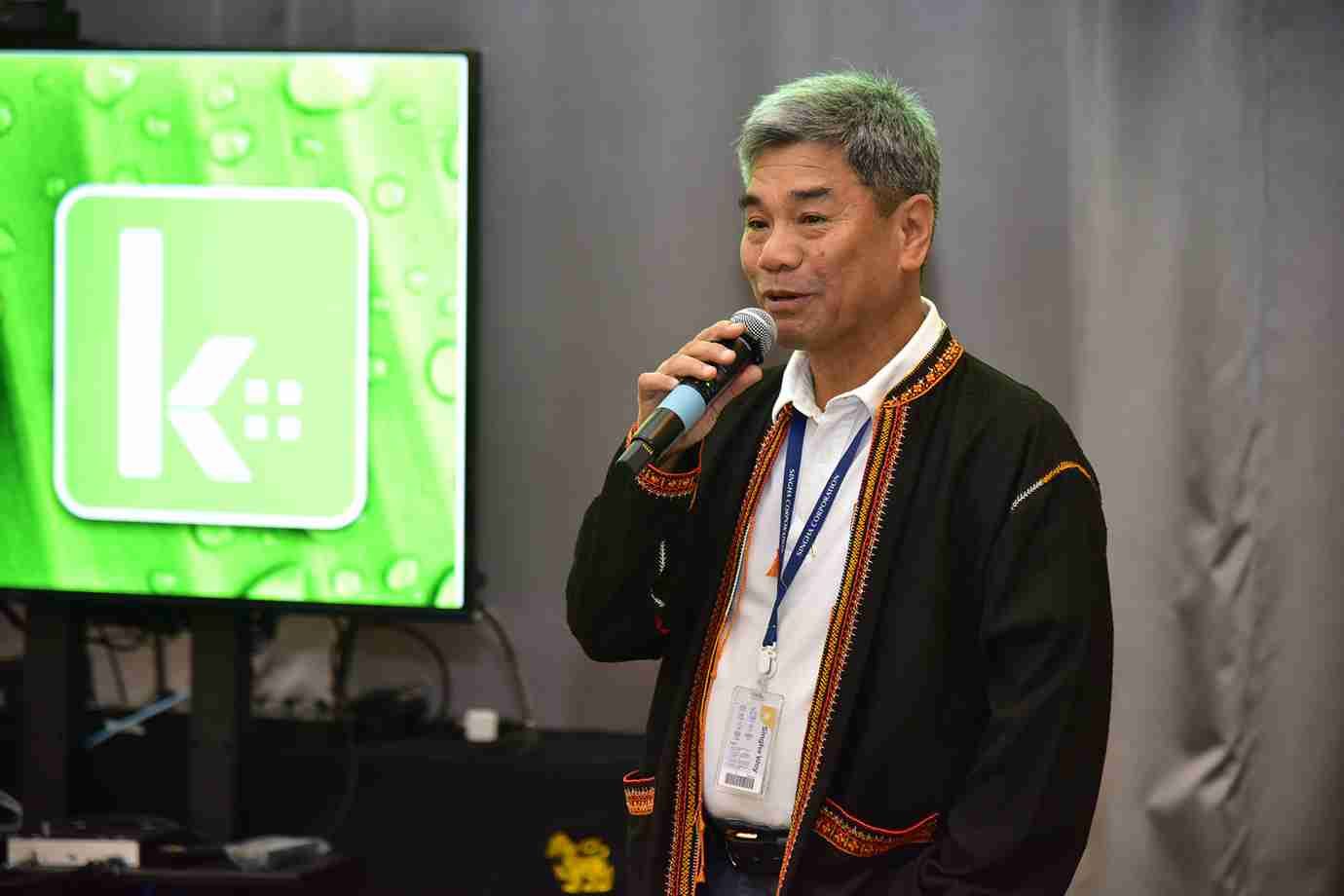 A man with grey hair in a traditional jacket speaks into a microphone, green logo screen visible.