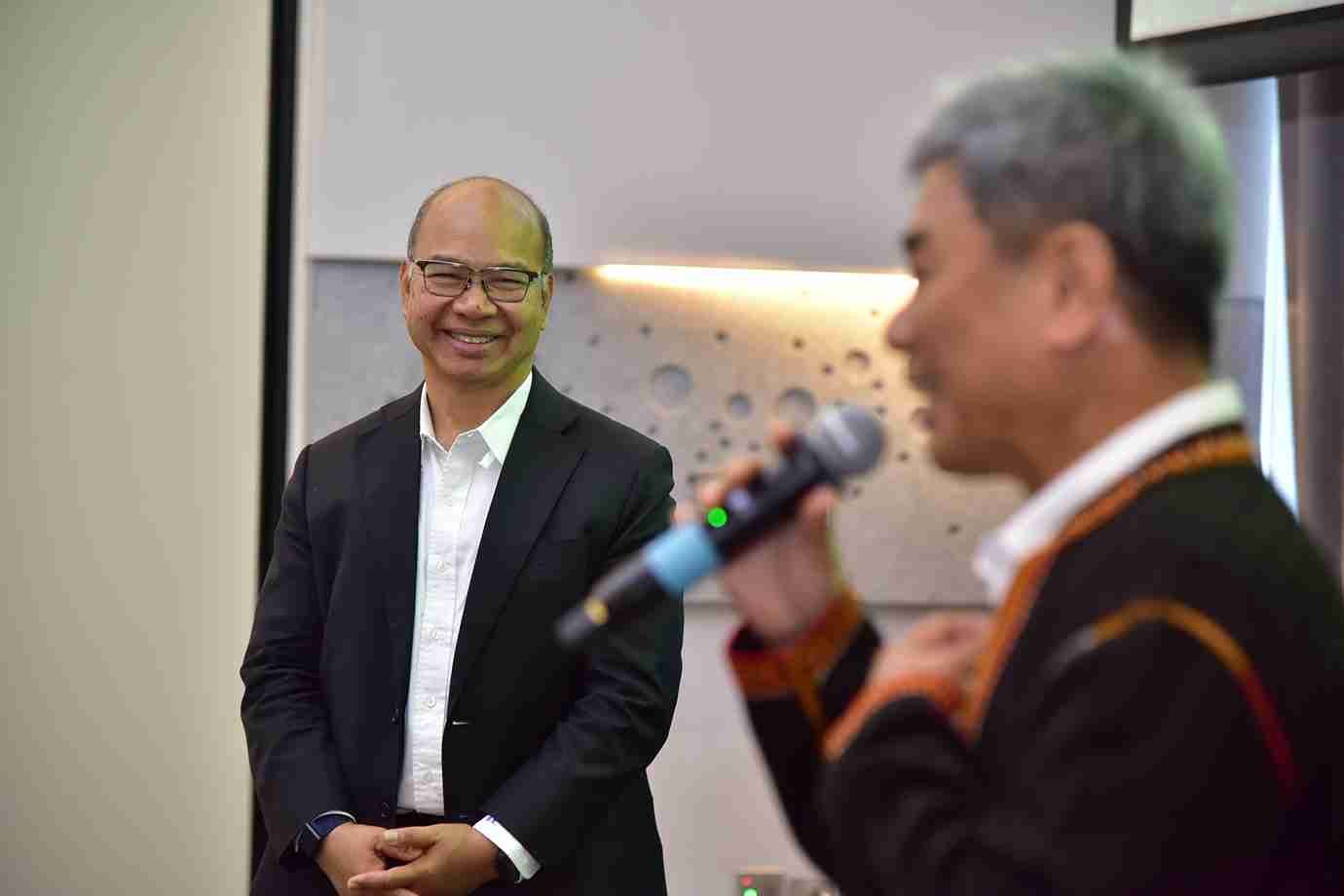 Smiling man in black suit watches another man speaking into a microphone.