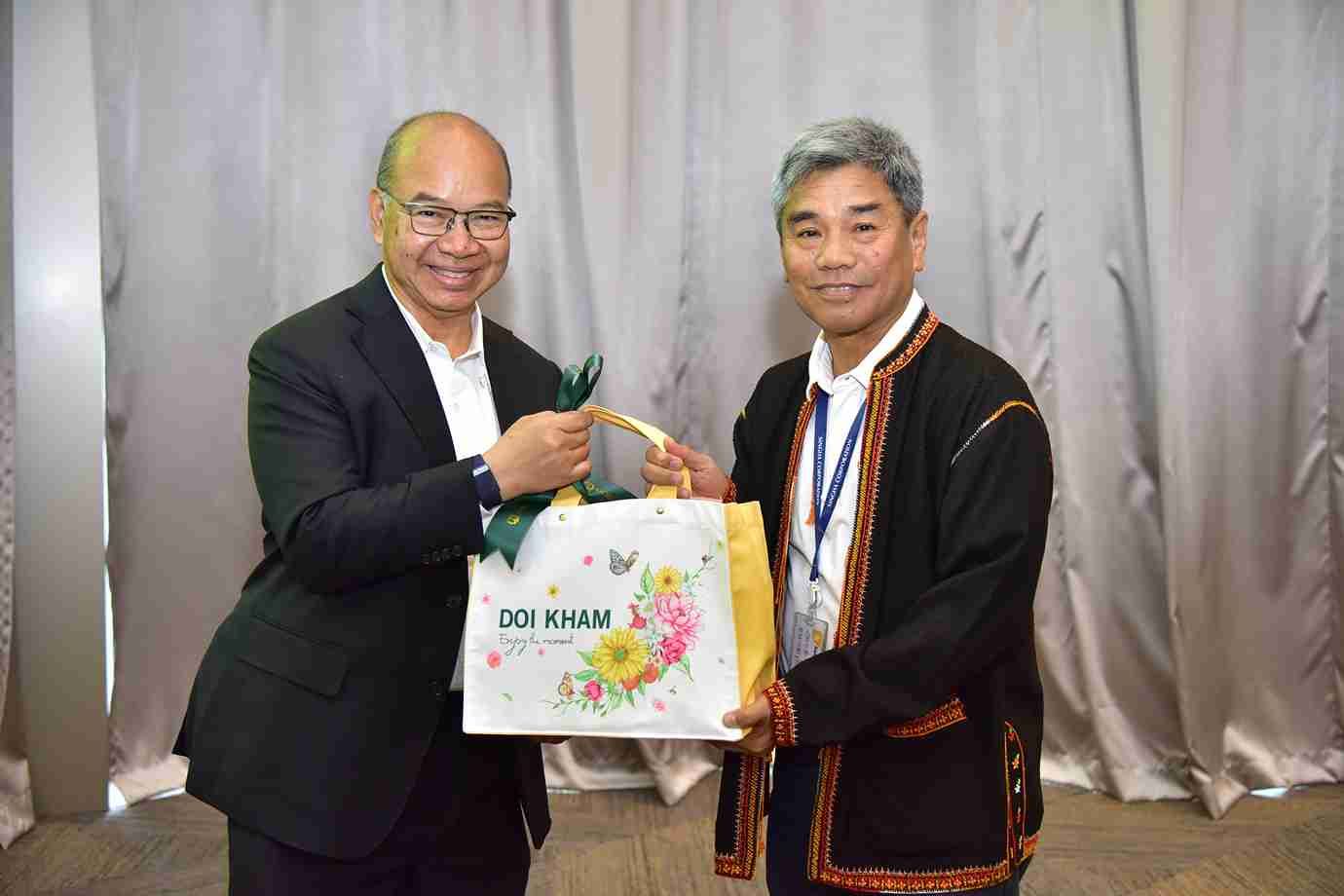 Two men exchange a colorful gift bag with 'DOI KHAM' printed on it.
