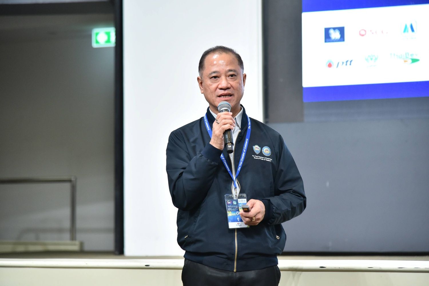 A man in a blue jacket speaks into a microphone, addressing an audience.