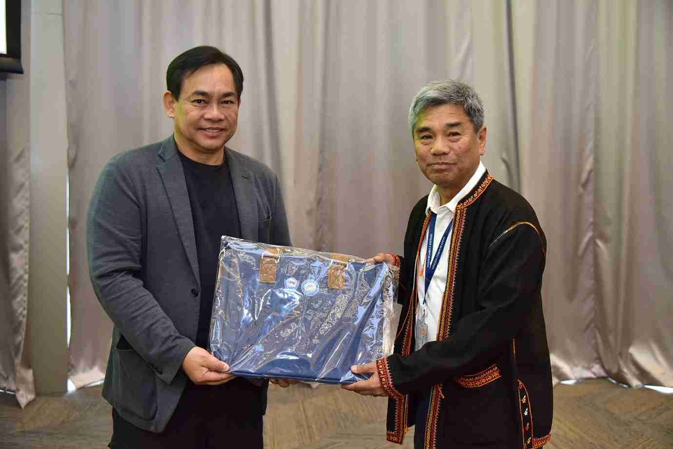 Two men exchanging a blue wrapped gift or award, smiling slightly.