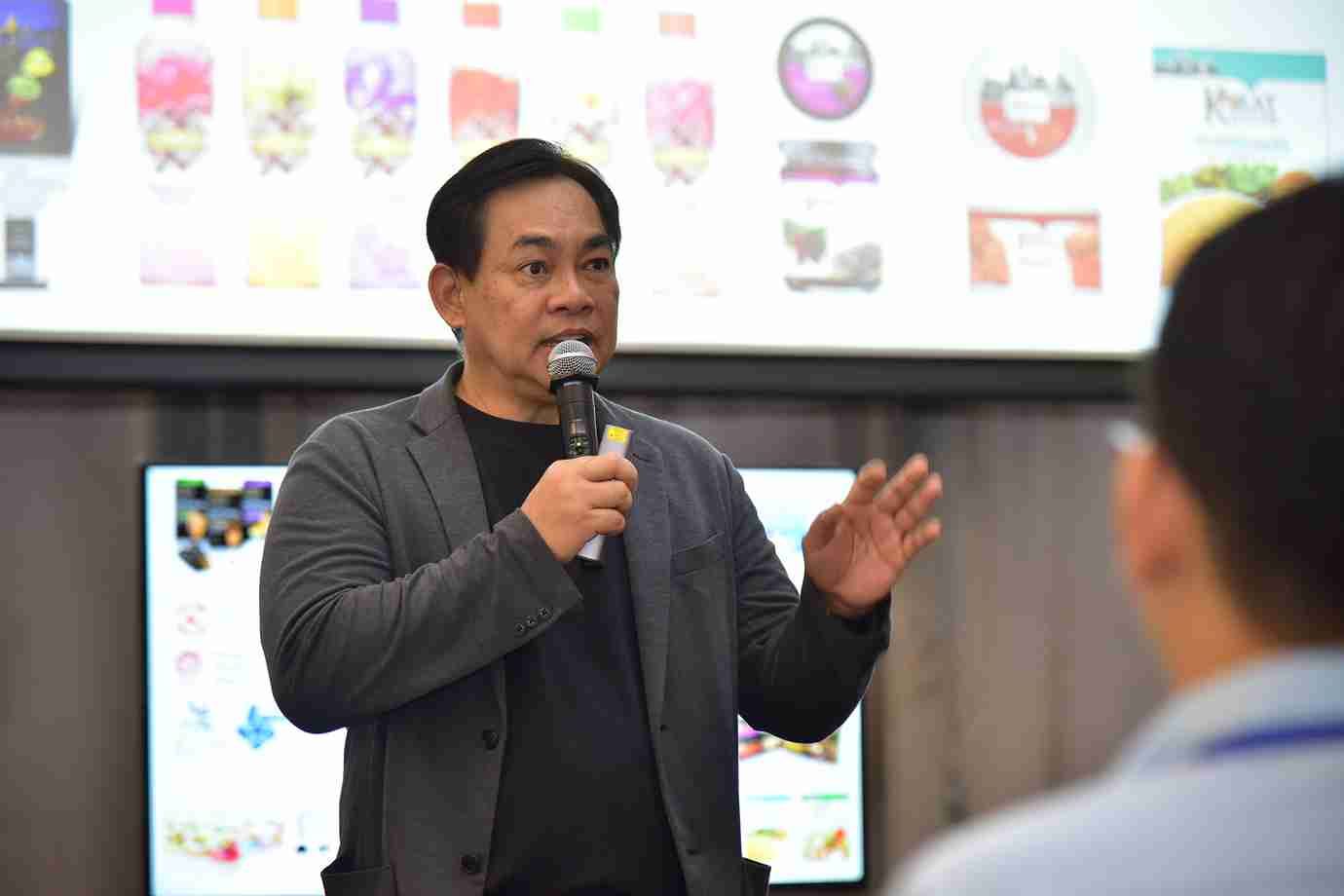 Man speaking into a microphone, gesturing, with product logos displayed on a screen behind him.