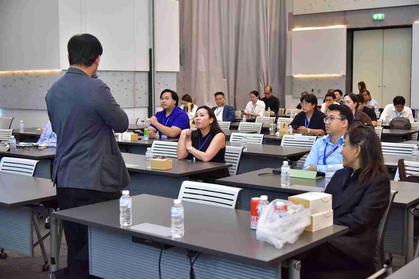 A speaker addresses an audience seated at tables in a brightly lit conference room.