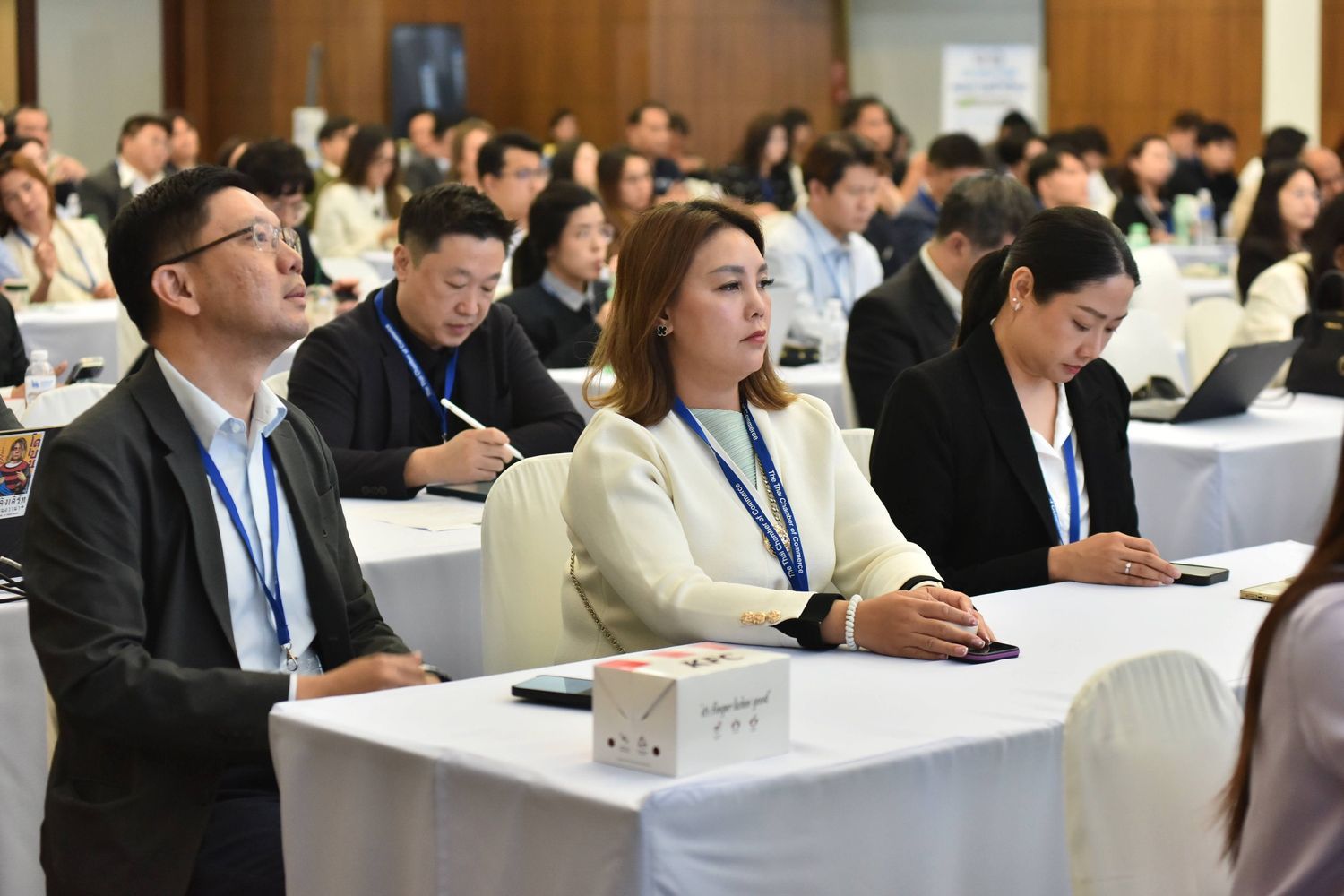 Conference attendees sit at tables, looking forward and listening attentively during a session.