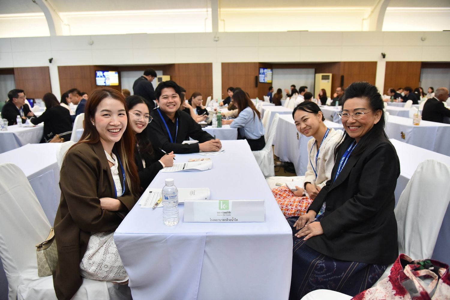 Six smiling attendees pose for a photo at a long white rectangular table in a conference room.