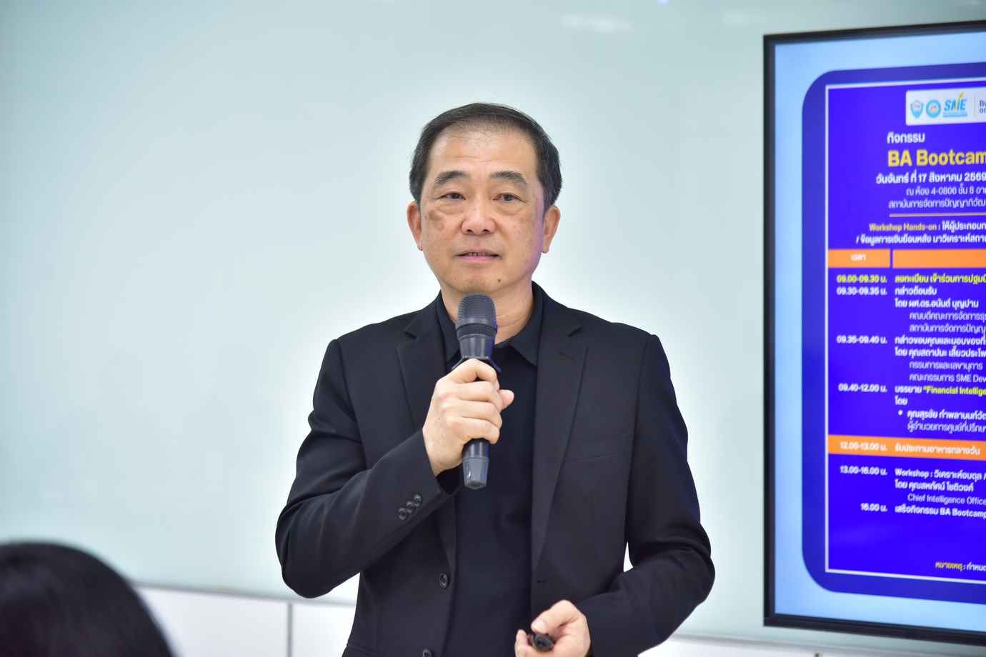 An Asian man in a dark suit presents into a microphone with a digital screen nearby.