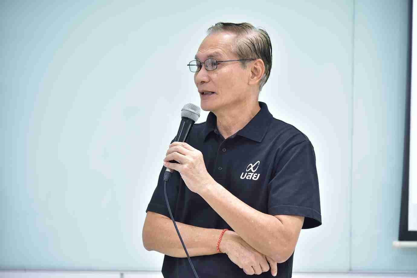 Man with glasses in dark polo shirt speaks into microphone, arms crossed.