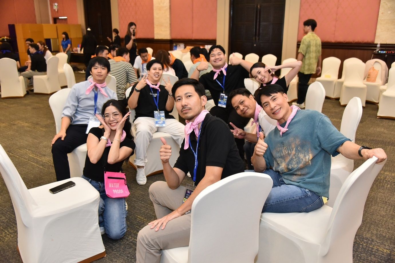 Smiling young adults in pink scarves pose with thumbs up in a large meeting room.