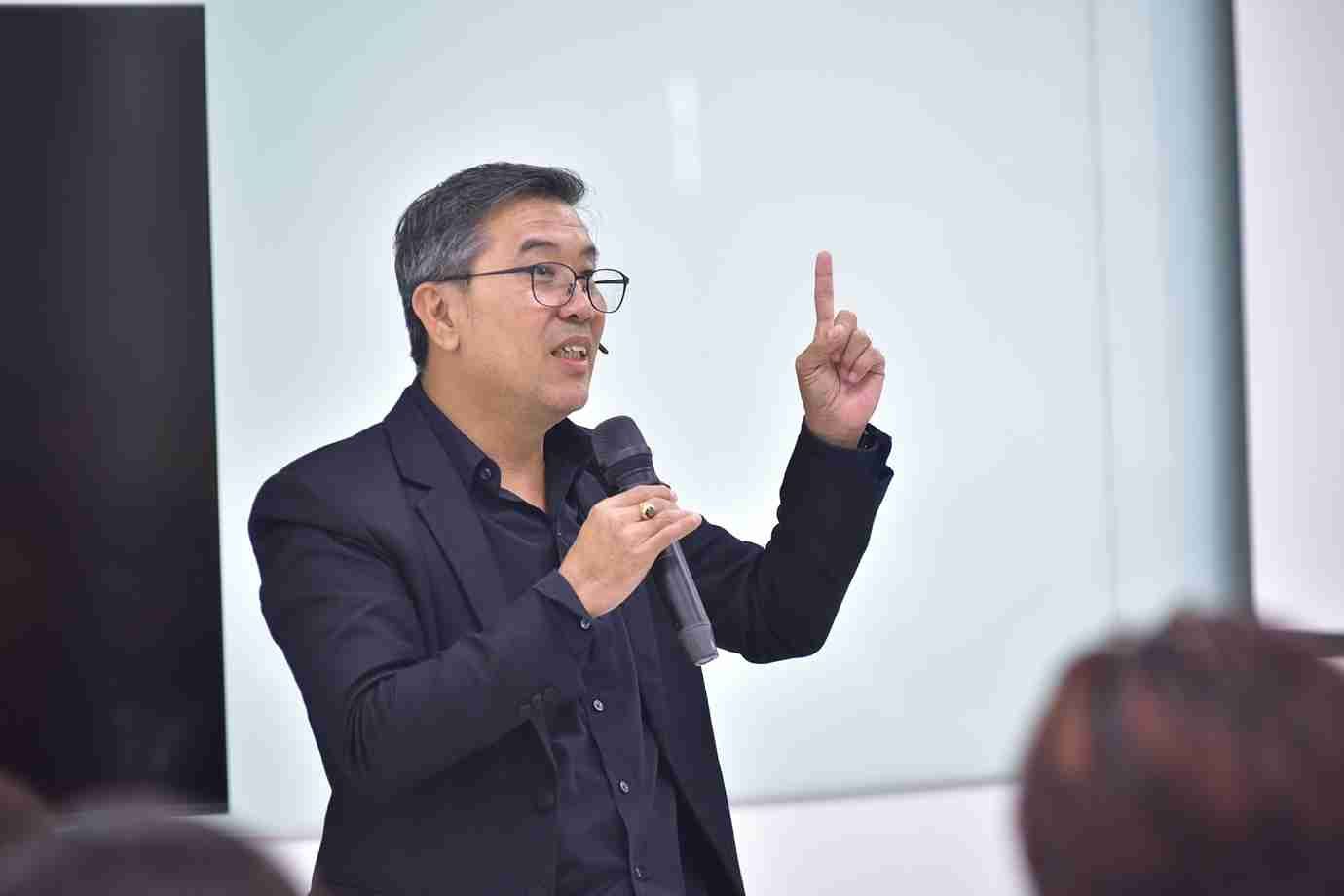 Man in glasses speaks into a microphone, gesturing with his index finger raised.