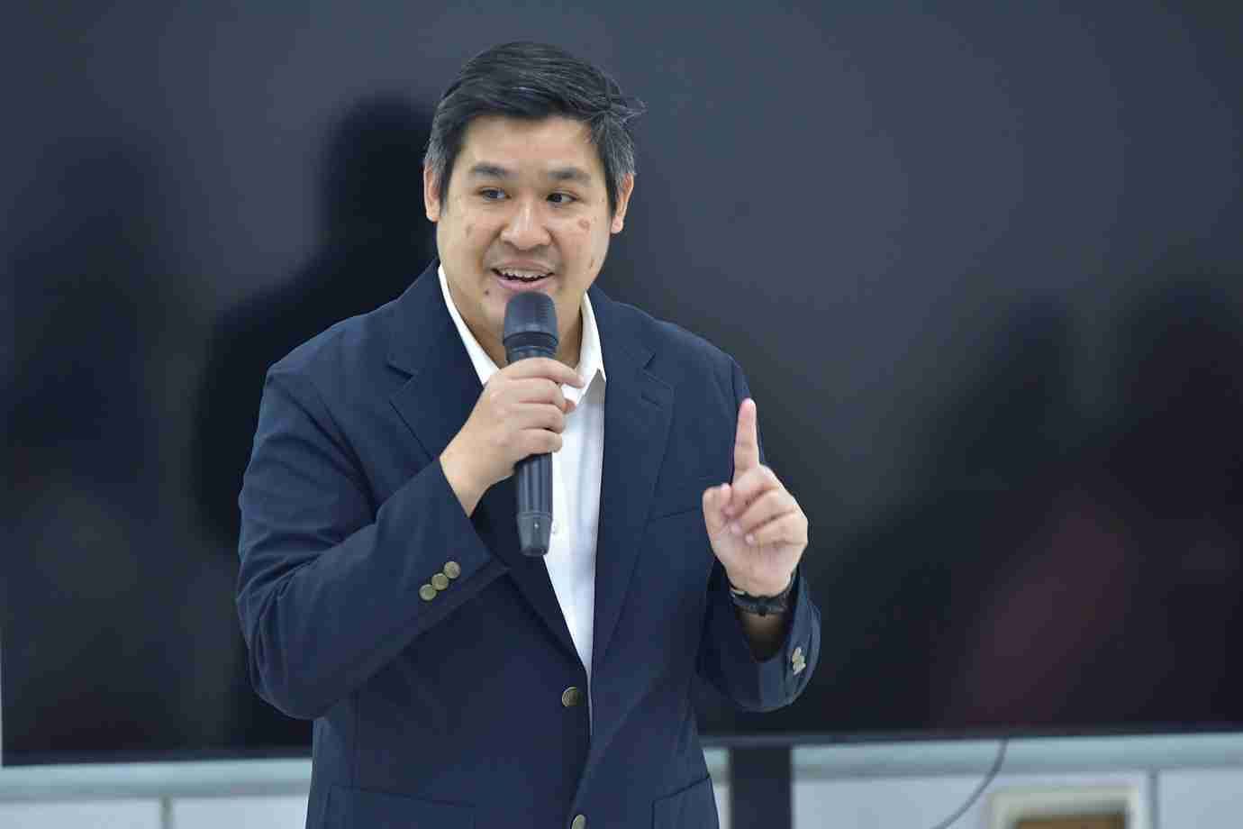 Man in dark suit jacket speaking into a microphone, gesturing with his finger.