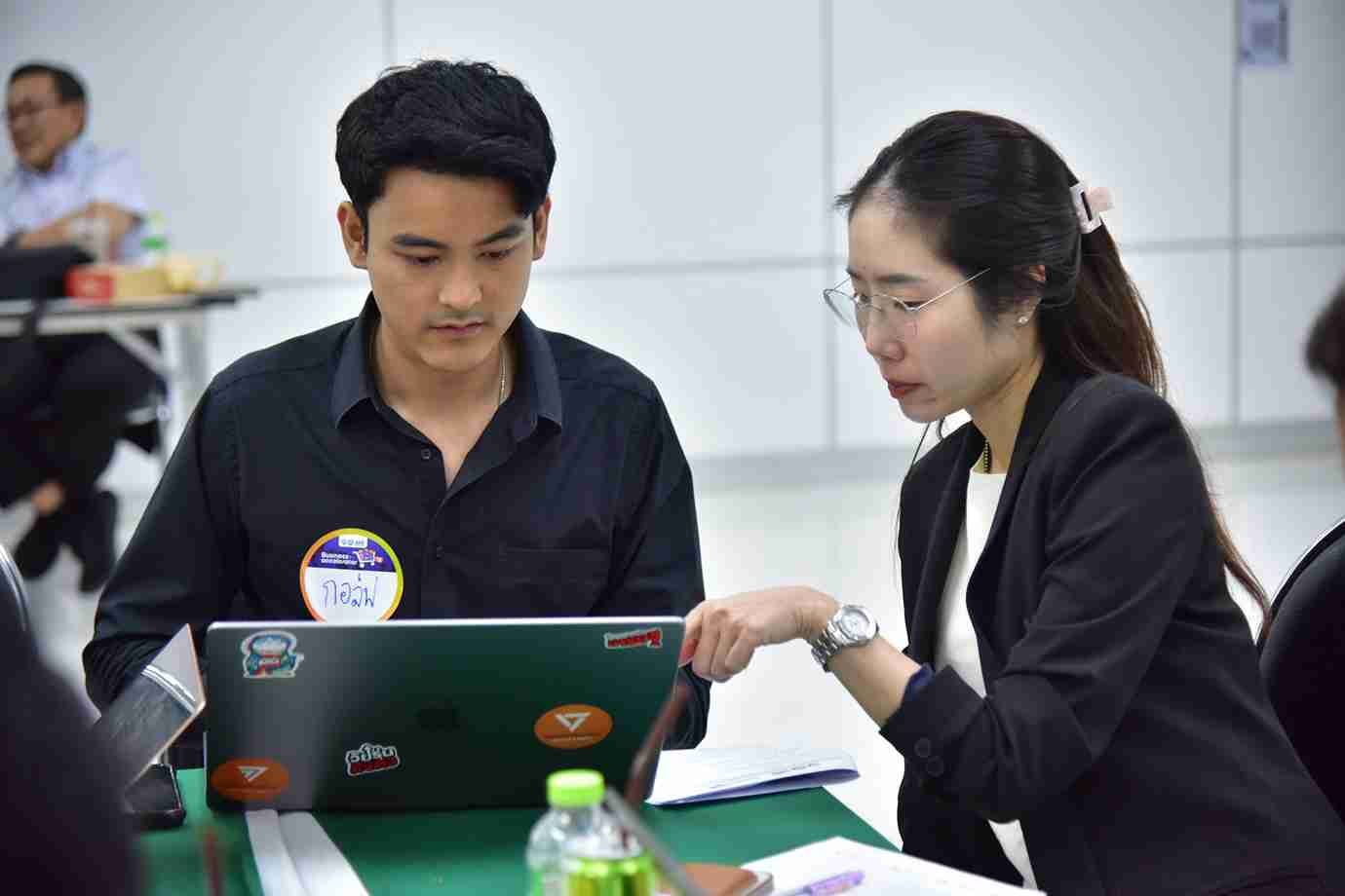 Two young professionals, a man and a woman, collaborate on a laptop in a meeting setting.