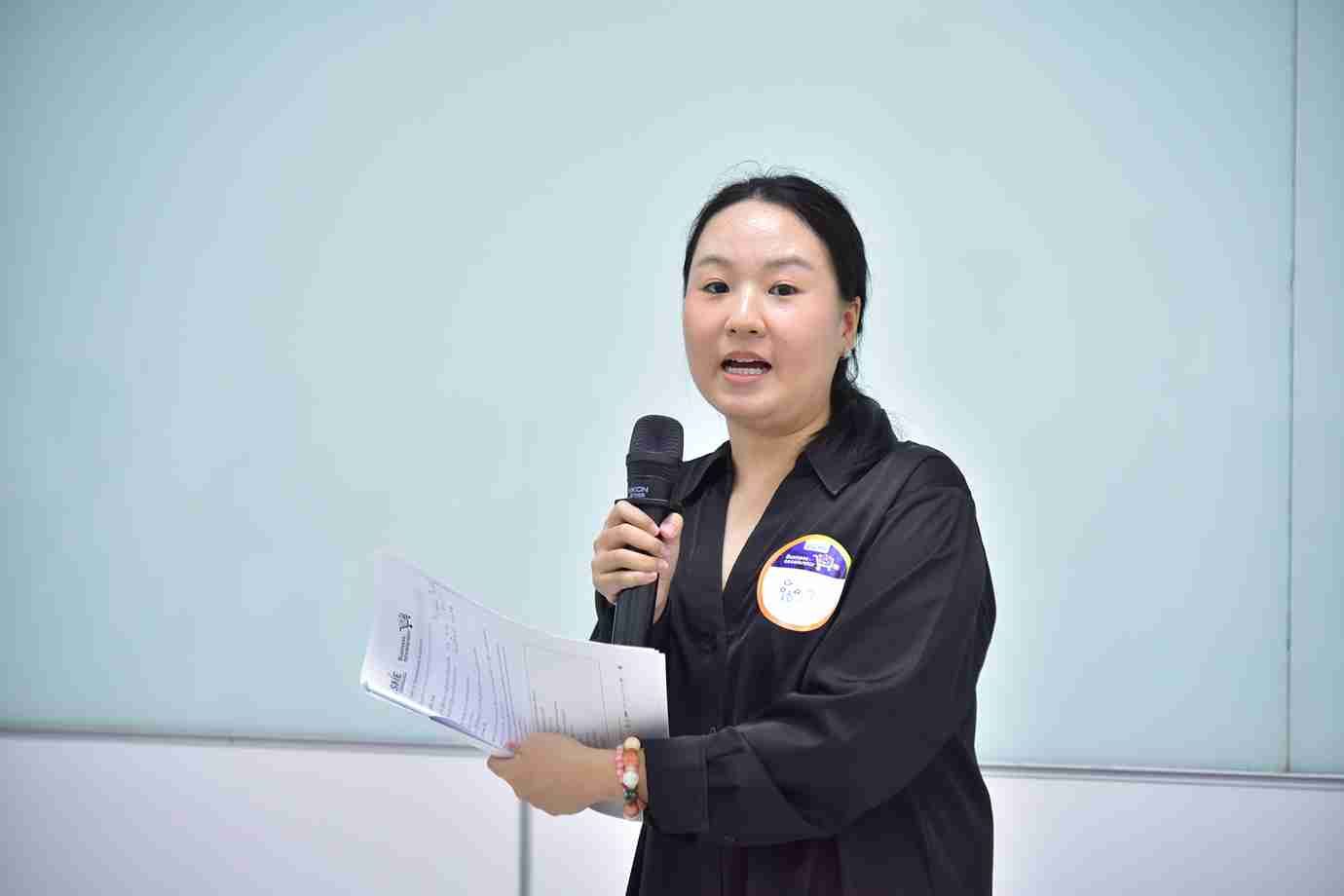 A woman in a dark shirt speaks into a microphone, holding papers.