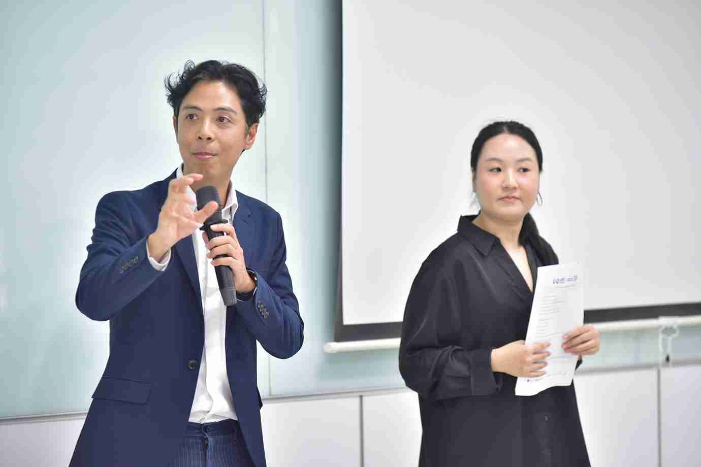 A male presenter speaks into a microphone next to a female colleague holding papers.