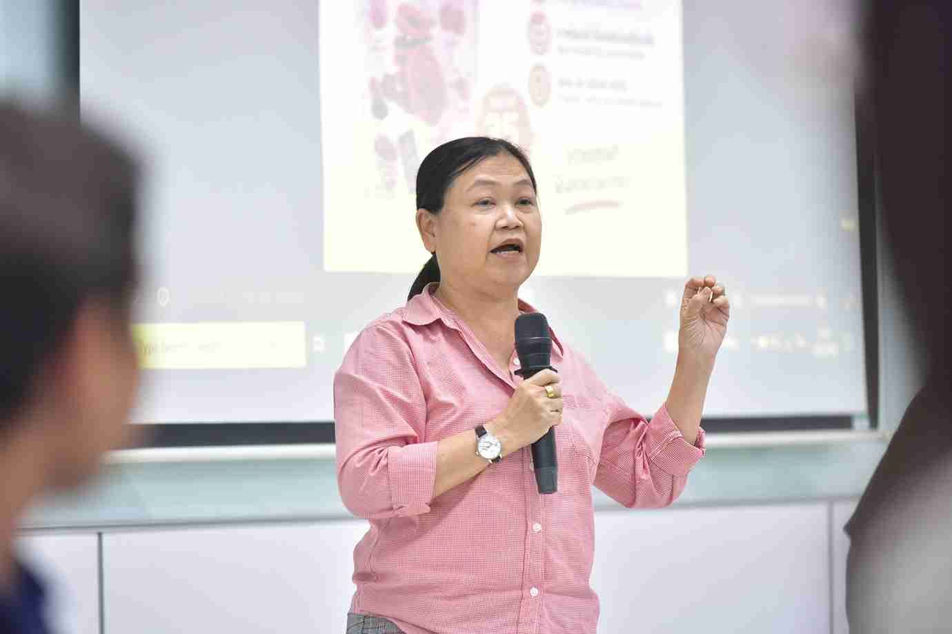 Woman in pink shirt speaks into microphone, gesturing, in front of a projector screen.
